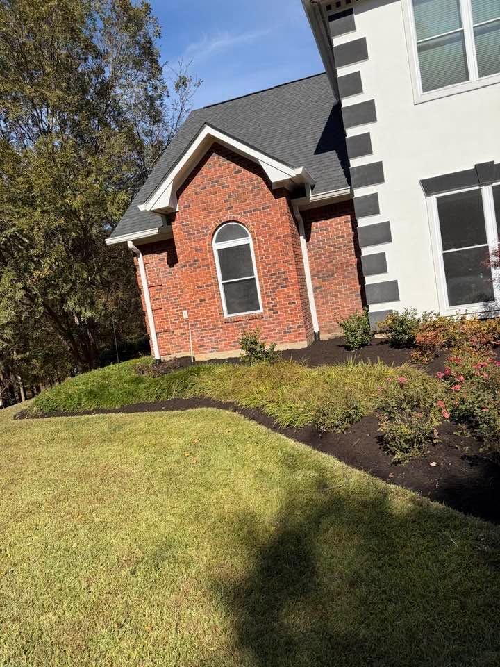 Brick house exterior with arched window, gray siding, lawn, and landscaped shrubs on a sunny day