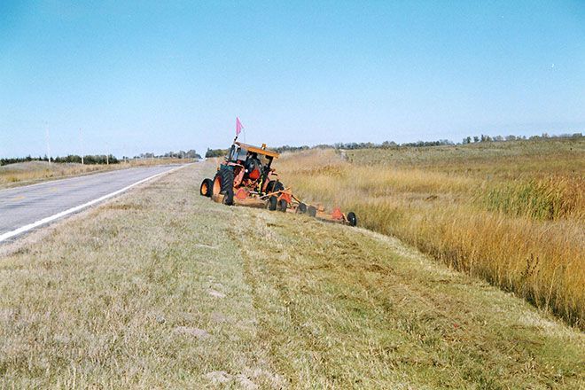 Roadside tractor mowing tall grass along a rural ditch under a clear blue sky