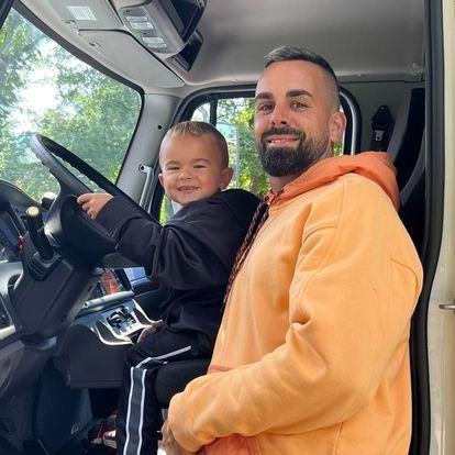 Man and child smiling in truck cab; child in driver's seat, man in orange hoodie.