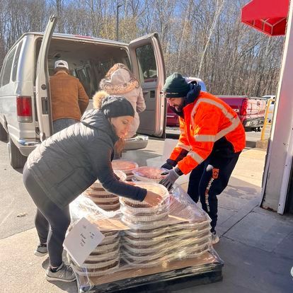 People unloading a van filled with packaged food pallets. A person in an orange vest assists. Sunny day.