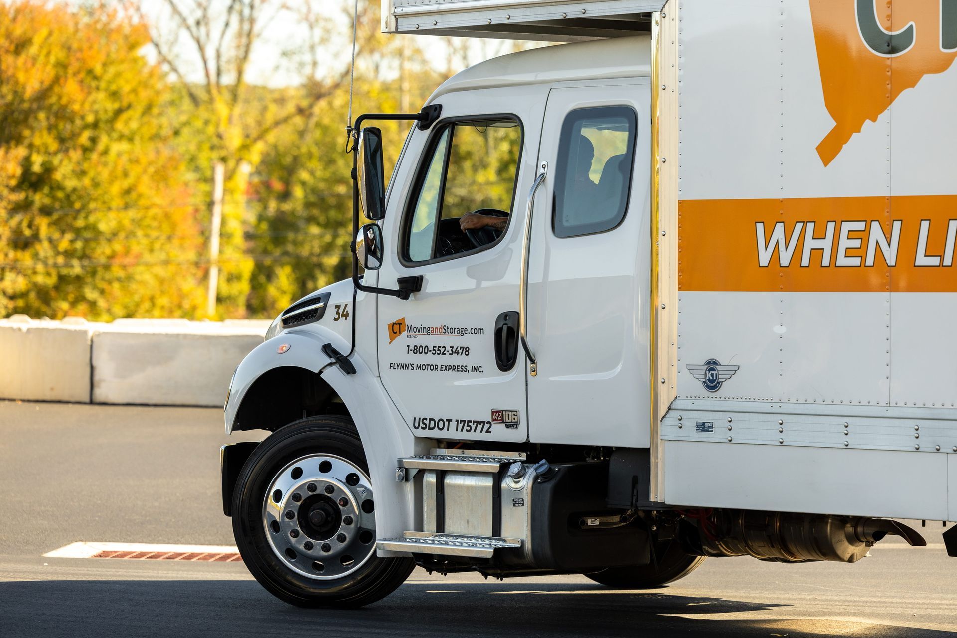 White moving truck with orange accents on asphalt. Driver visible in the cab.