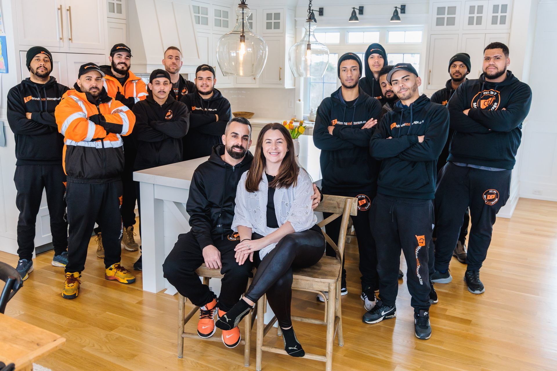 Group of people in black and orange attire, pose in a home with wooden floors, centered around two seated individuals.