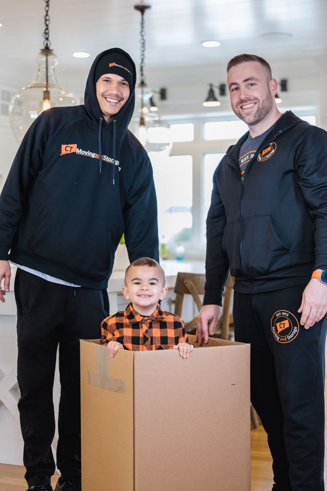 Two men and a child smiling, the child in a cardboard box indoors.