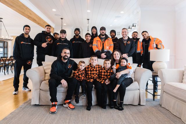 Group of people, including children, pose for a photo in a living room. Some wear orange and black work attire.