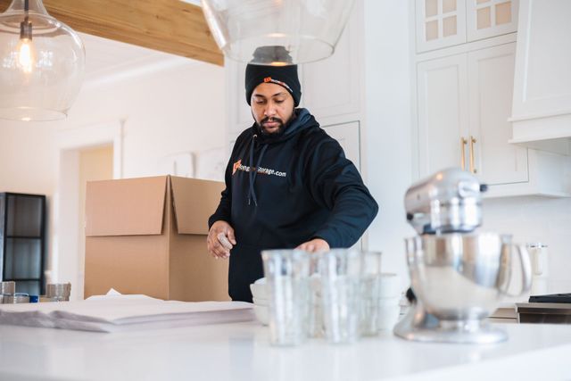 Man in a black hoodie and beanie packing kitchen items, surrounded by boxes and glassware on a white countertop.