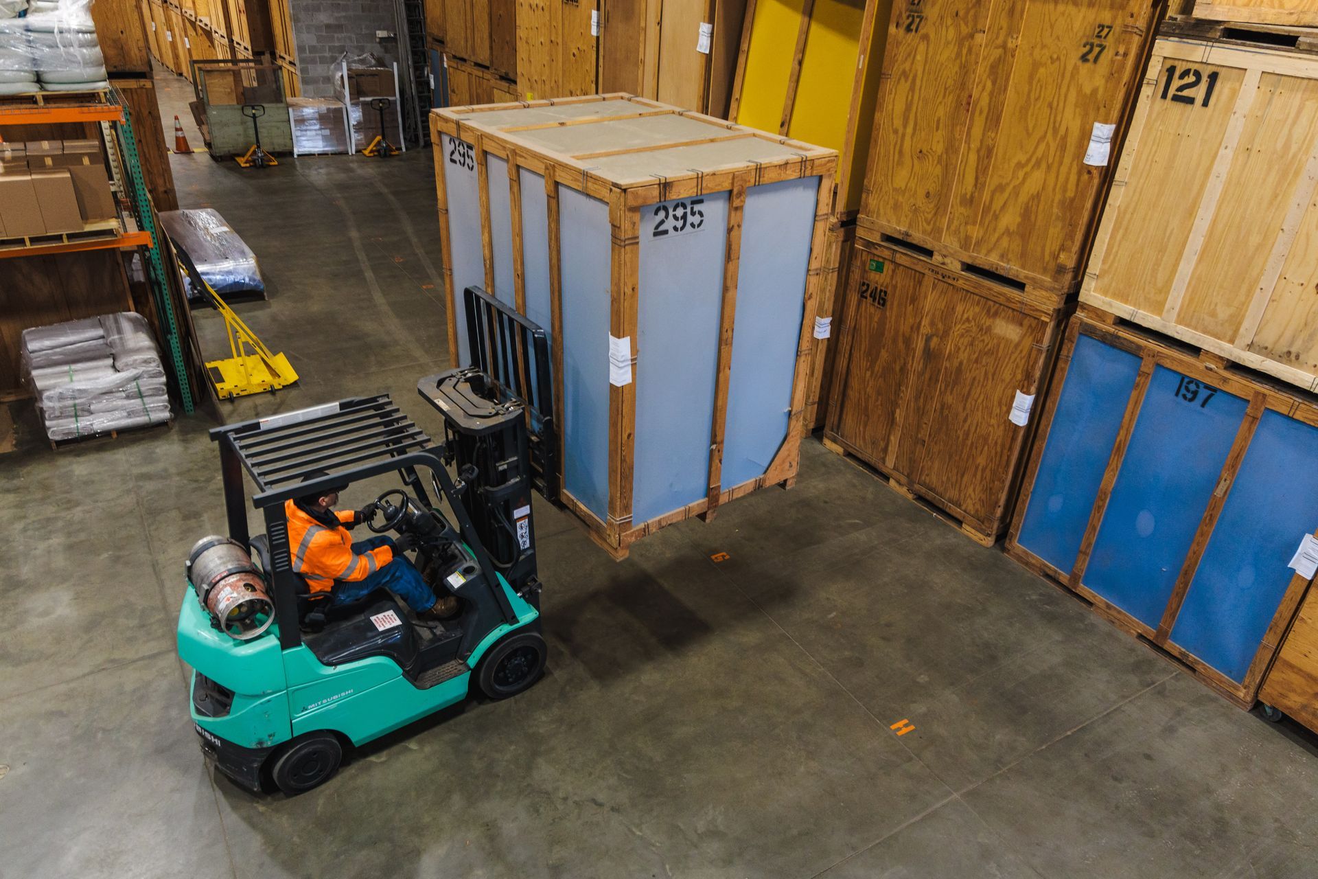Forklift lifts a large wooden crate in a warehouse, near other crates.