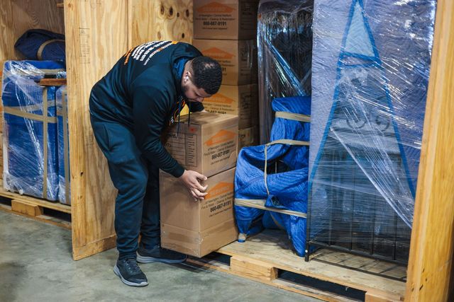 Man packing a cardboard box in a wooden storage unit, surrounded by wrapped furniture and other boxes.