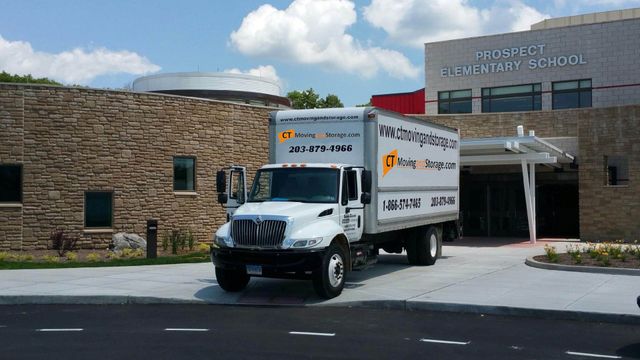 White moving truck in front of a school with a stone facade, under a sunny sky.