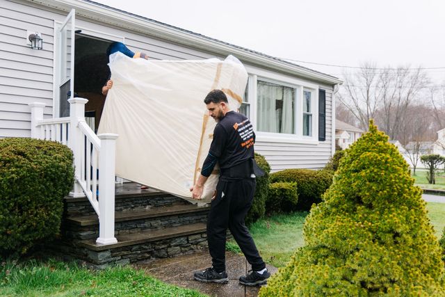 Two movers carrying a mattress out of a house's doorway.
