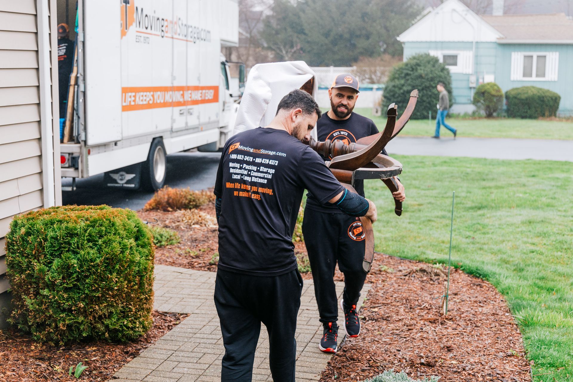 Two movers carrying furniture from a house towards a moving truck. Exterior setting.
