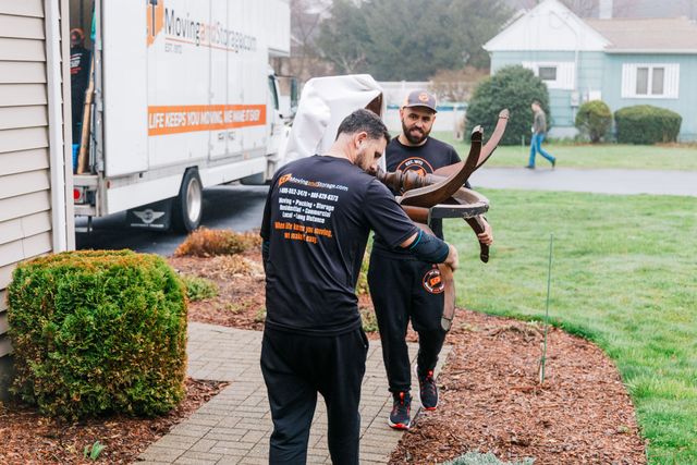 Two movers carrying furniture from a house towards a moving truck. Exterior setting.