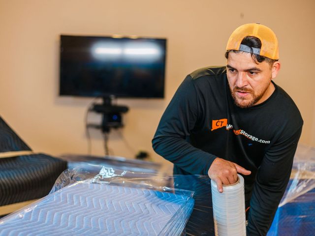 Man rolls packing material, near wrapped furniture and a TV. Indoors, neutral color setting.