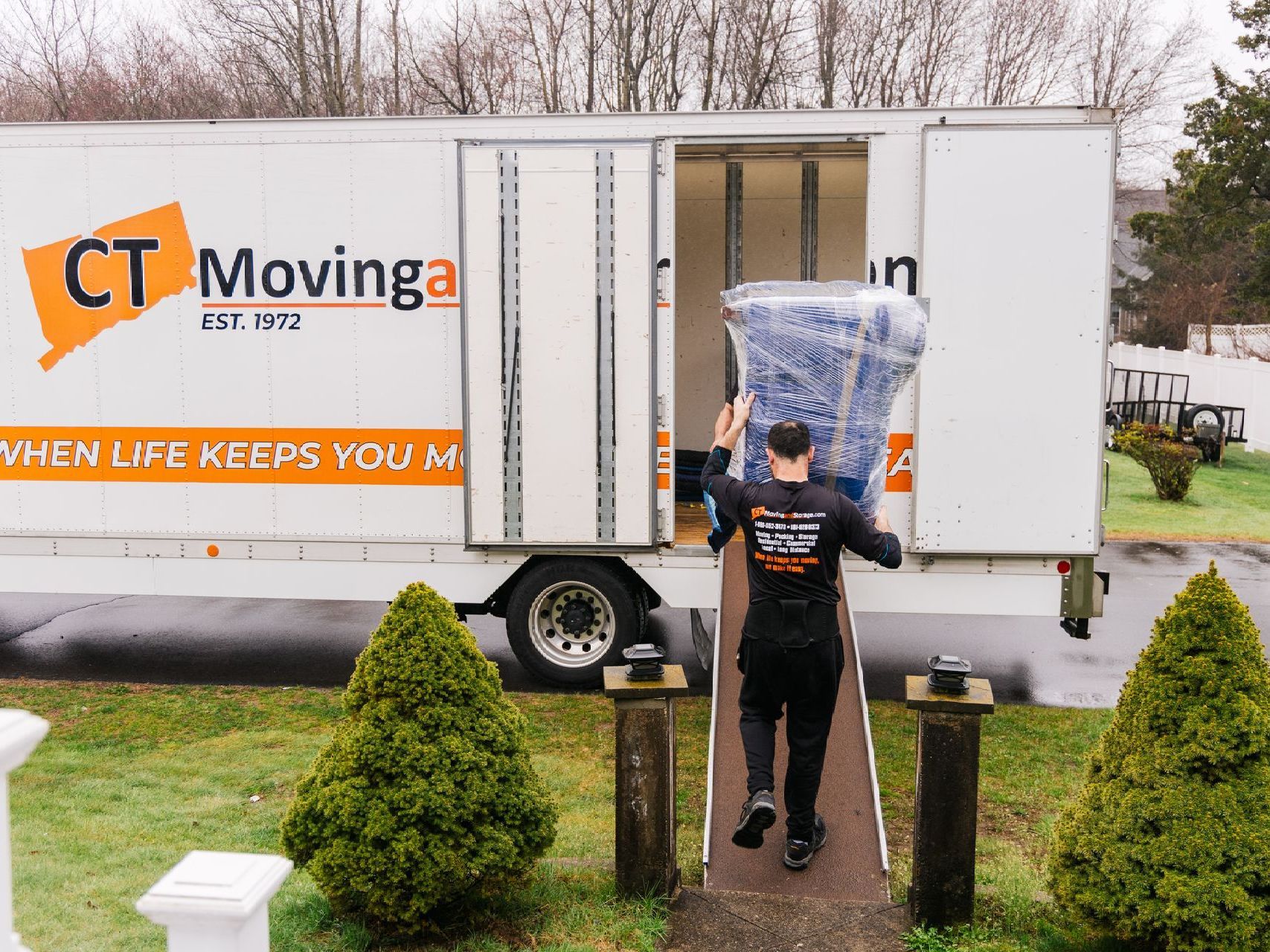 Man carrying wrapped furniture into a CT Moving truck. The truck is parked in a driveway in front of a house.