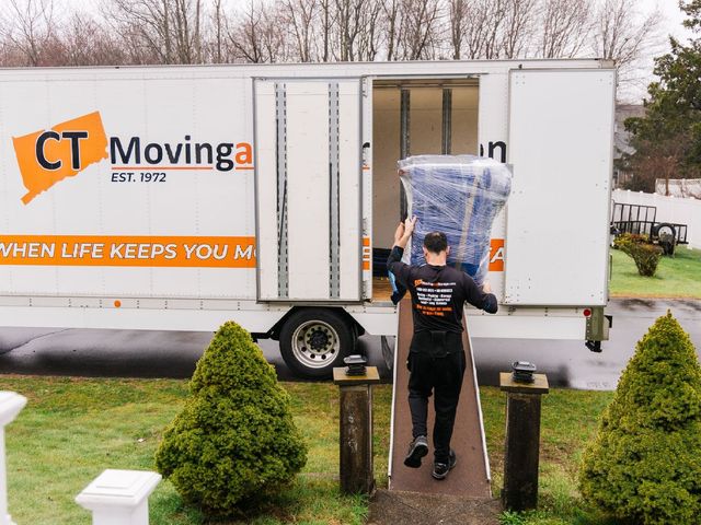 Man carrying wrapped furniture into a CT Moving truck. The truck is parked in a driveway in front of a house.