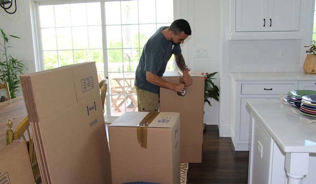 Man taping a cardboard box in a kitchen with large windows and a white countertop.