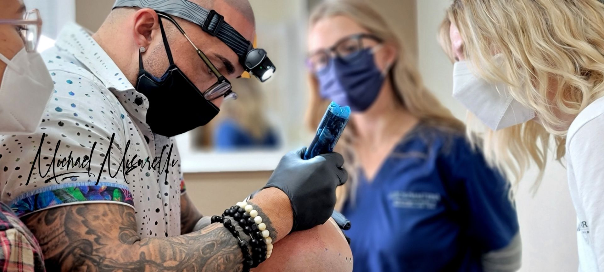 A person wearing a headlamp and mask performs a procedure on a person's head with medical staff present.