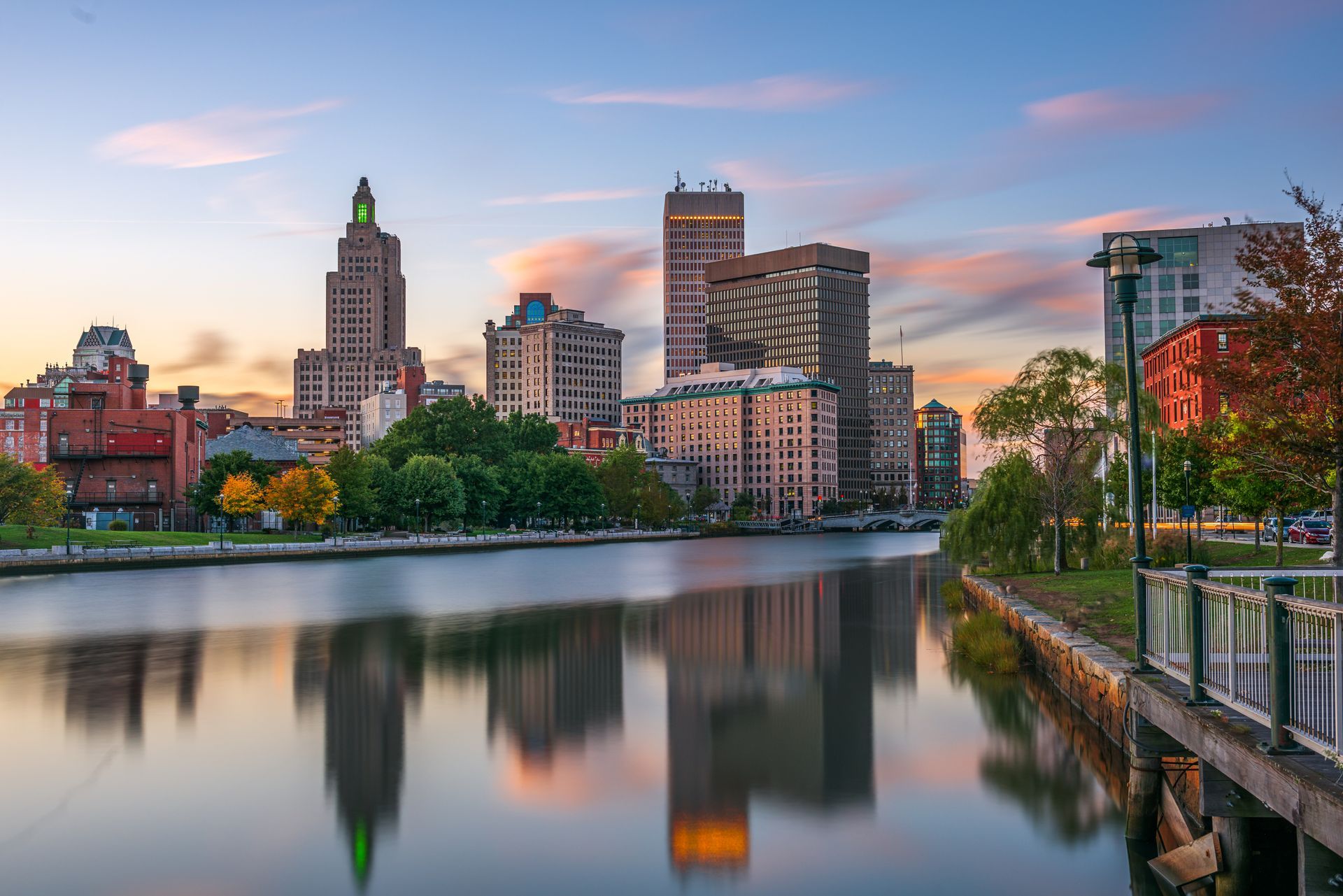 City skyline at sunset reflected in a calm river, with trees and a waterfront walkway in the foreground