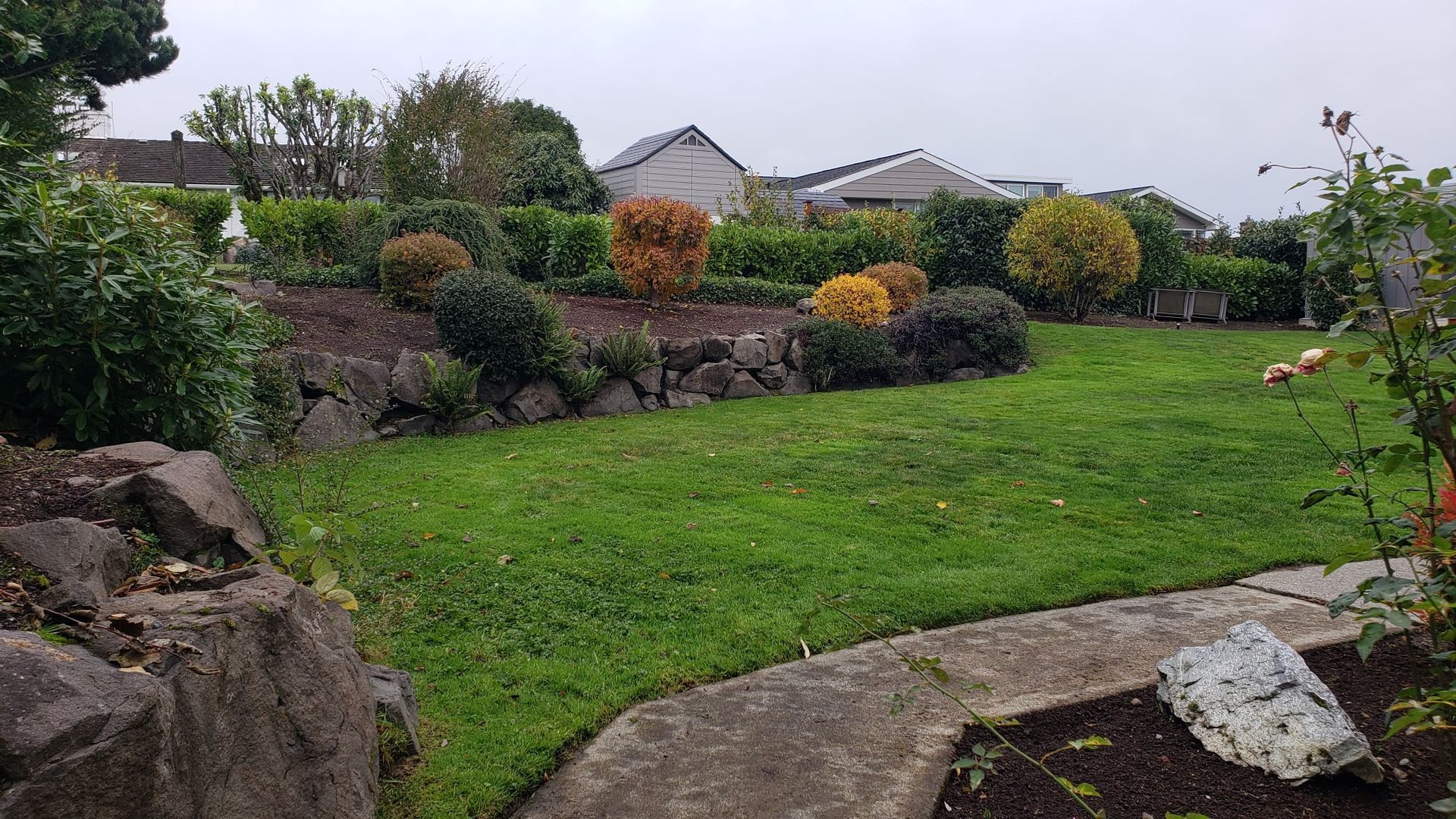Green lawn with rock border and tiered garden beds filled with various shrubs and trees.