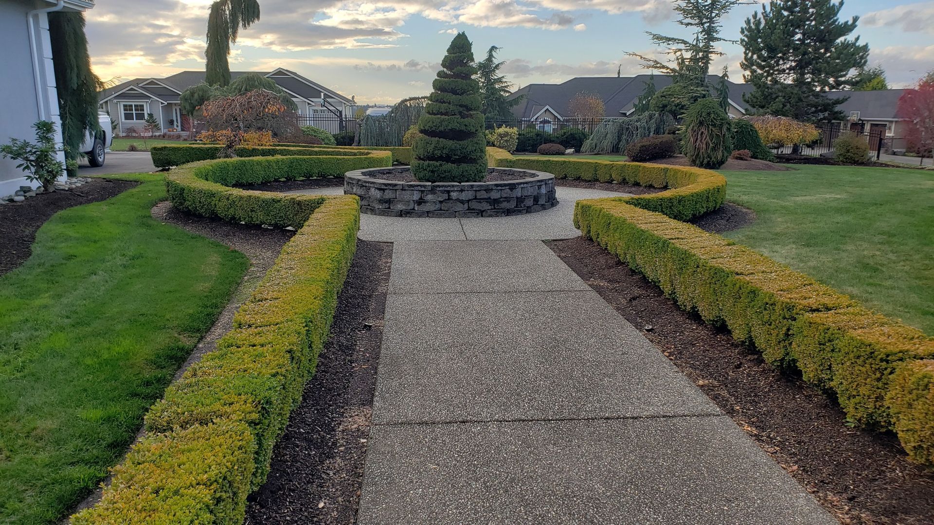 Stone path leads to a tiered topiary in a garden bed with curved hedges and surrounding houses under a cloudy sky.