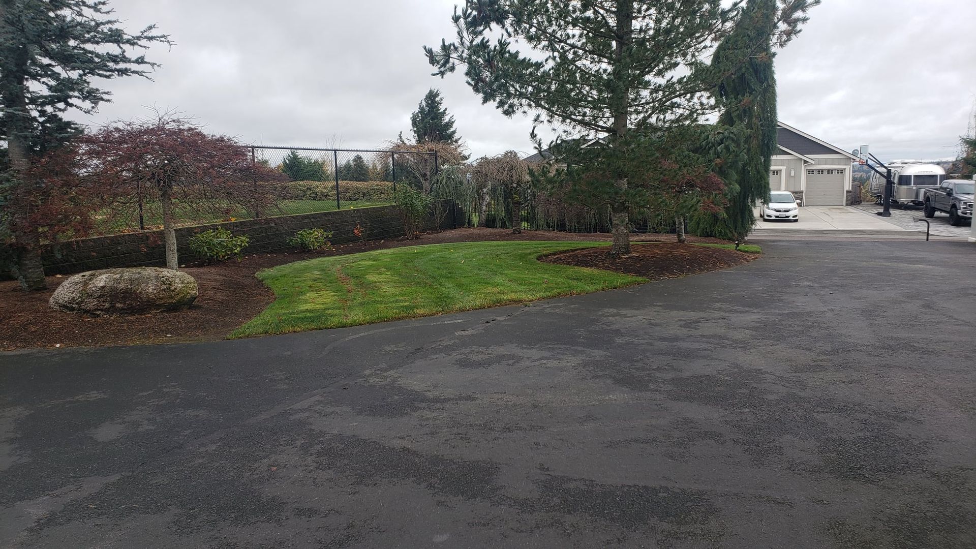 Asphalt driveway leads to a house with a landscaped garden on an overcast day.