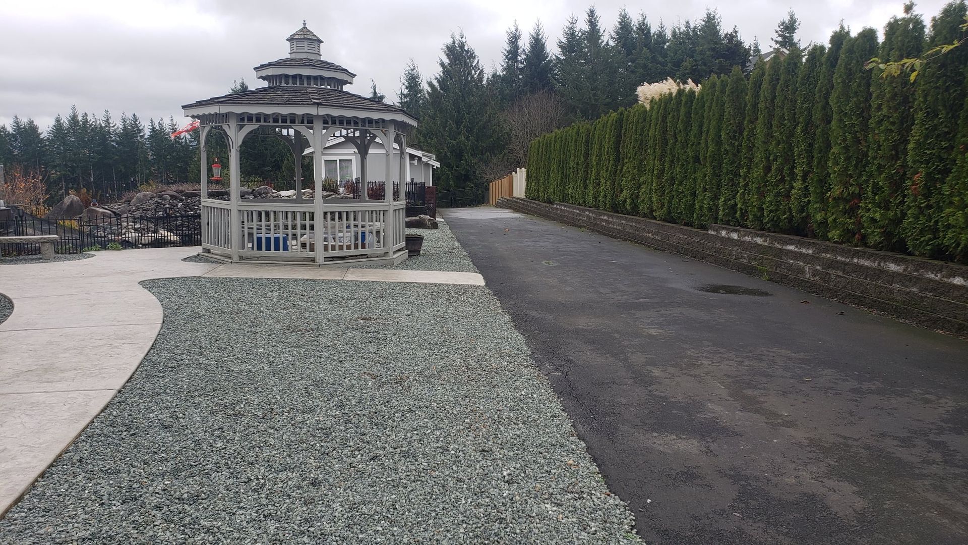 Gazebo with gravel path and asphalt driveway, bordered by green hedge. Cloudy day.