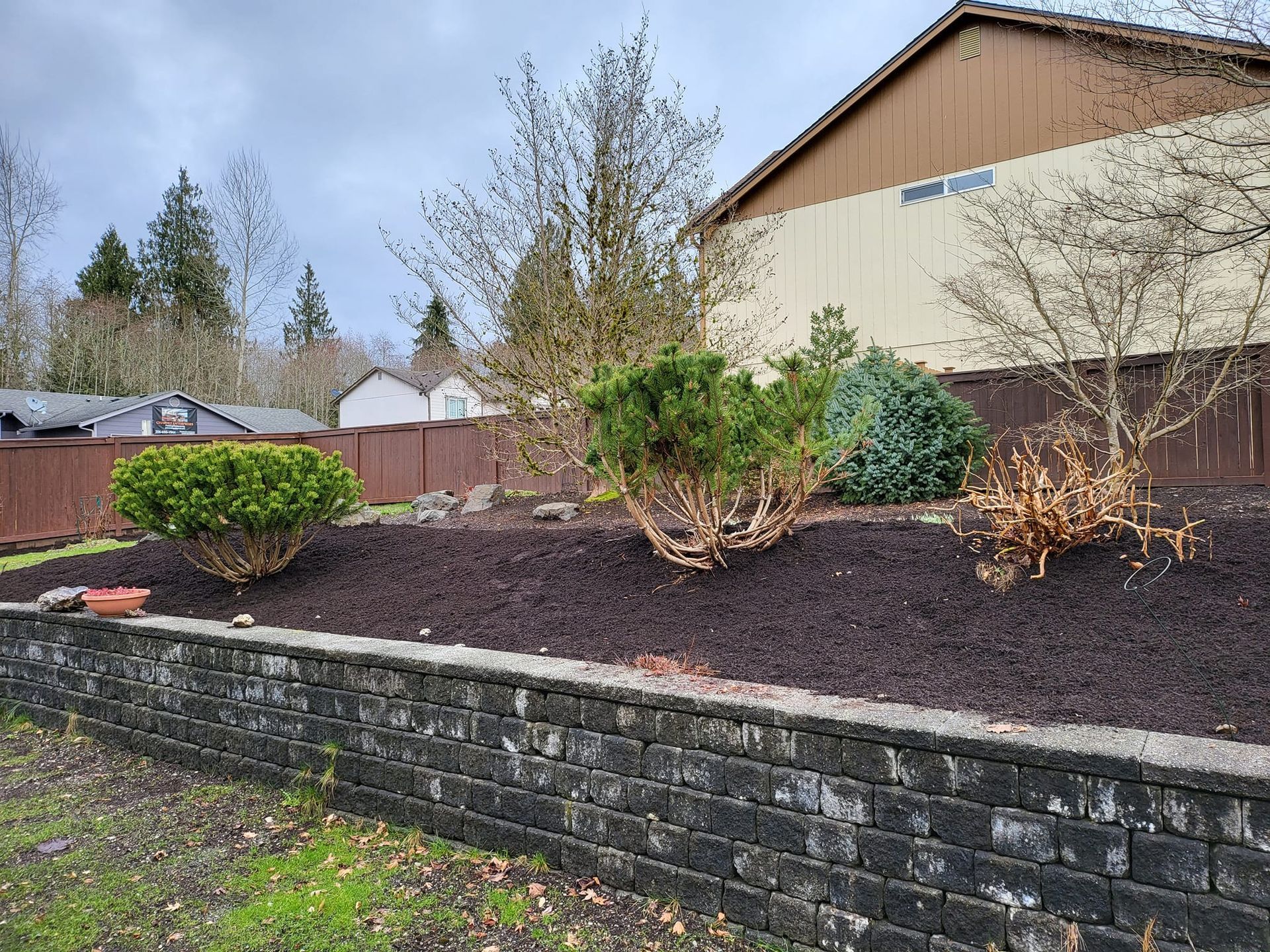 Stone retaining wall with a landscaped bed of mulch and shrubs in front of a house.