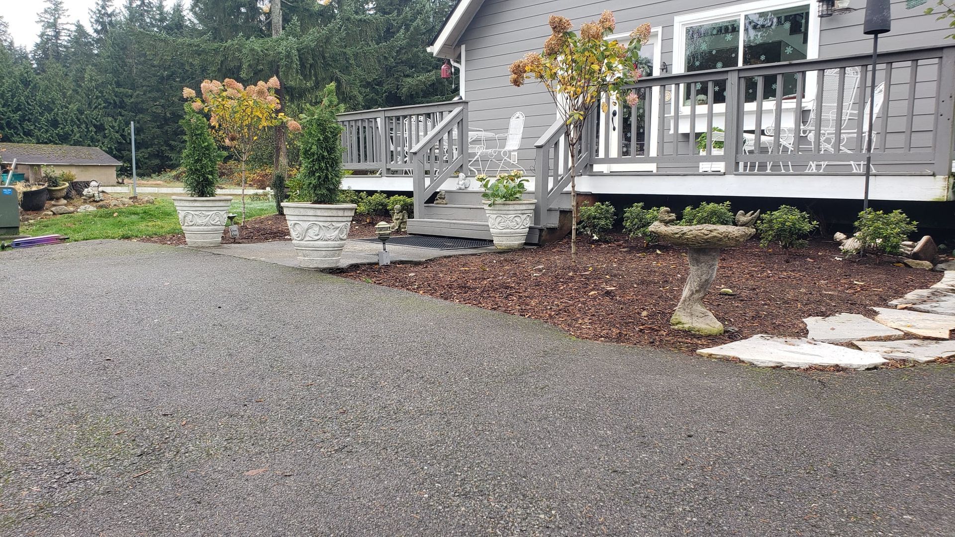 Gravel driveway leads to house with deck and gray siding, potted plants, and mulched landscaping.