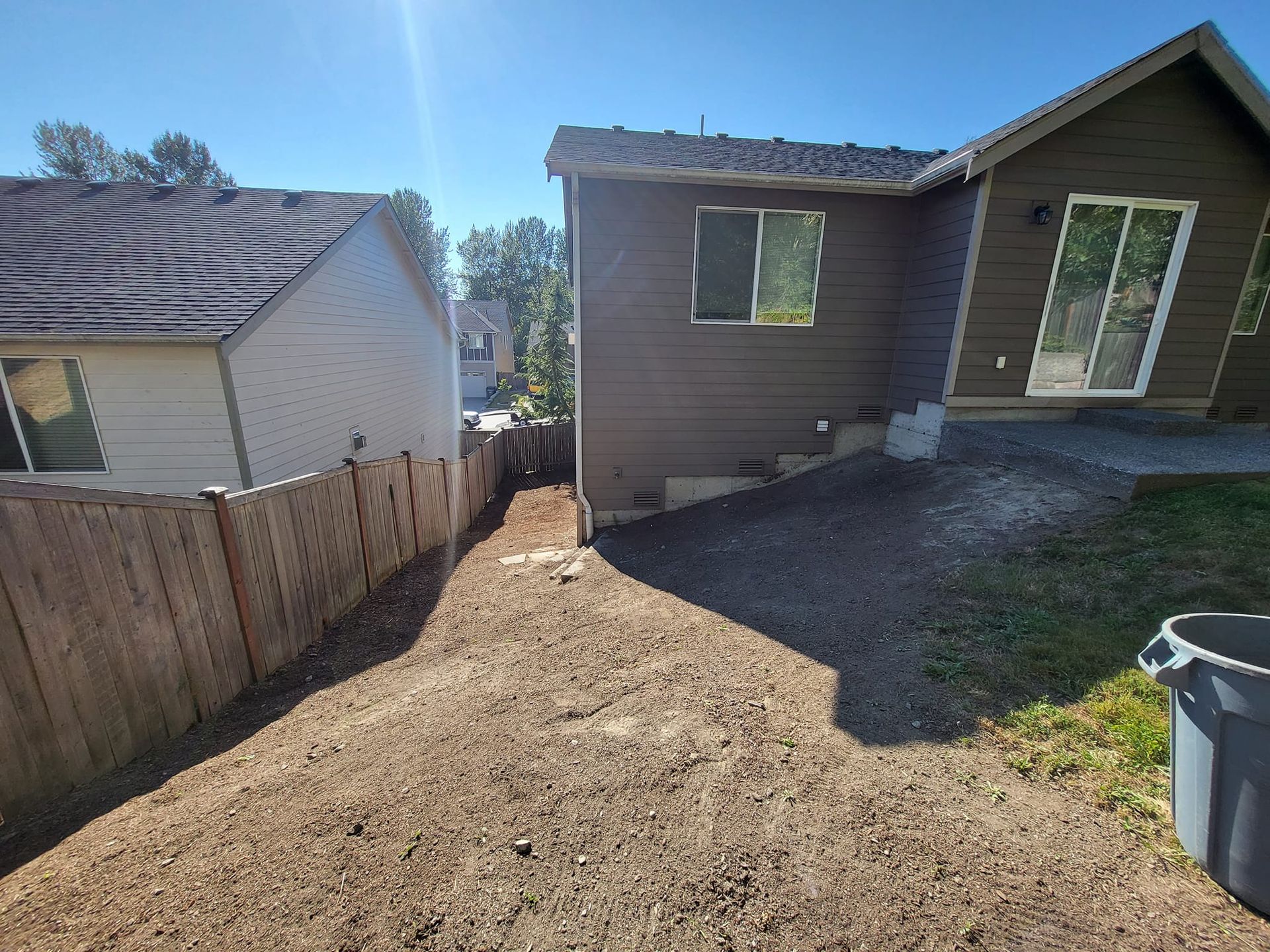 Backyard dirt path between a brown house and a wooden fence on a sunny day.