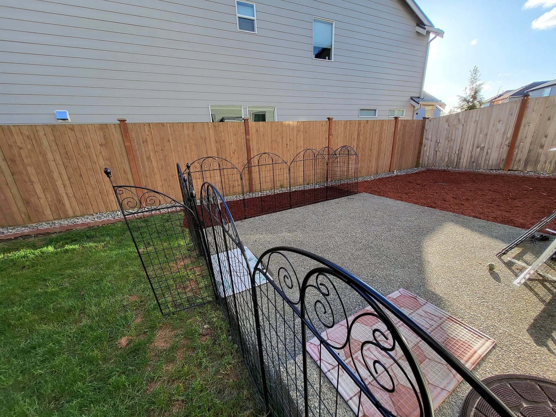 Backyard with a gravel area, red mulch, and a decorative black metal fence near a wooden fence and building.