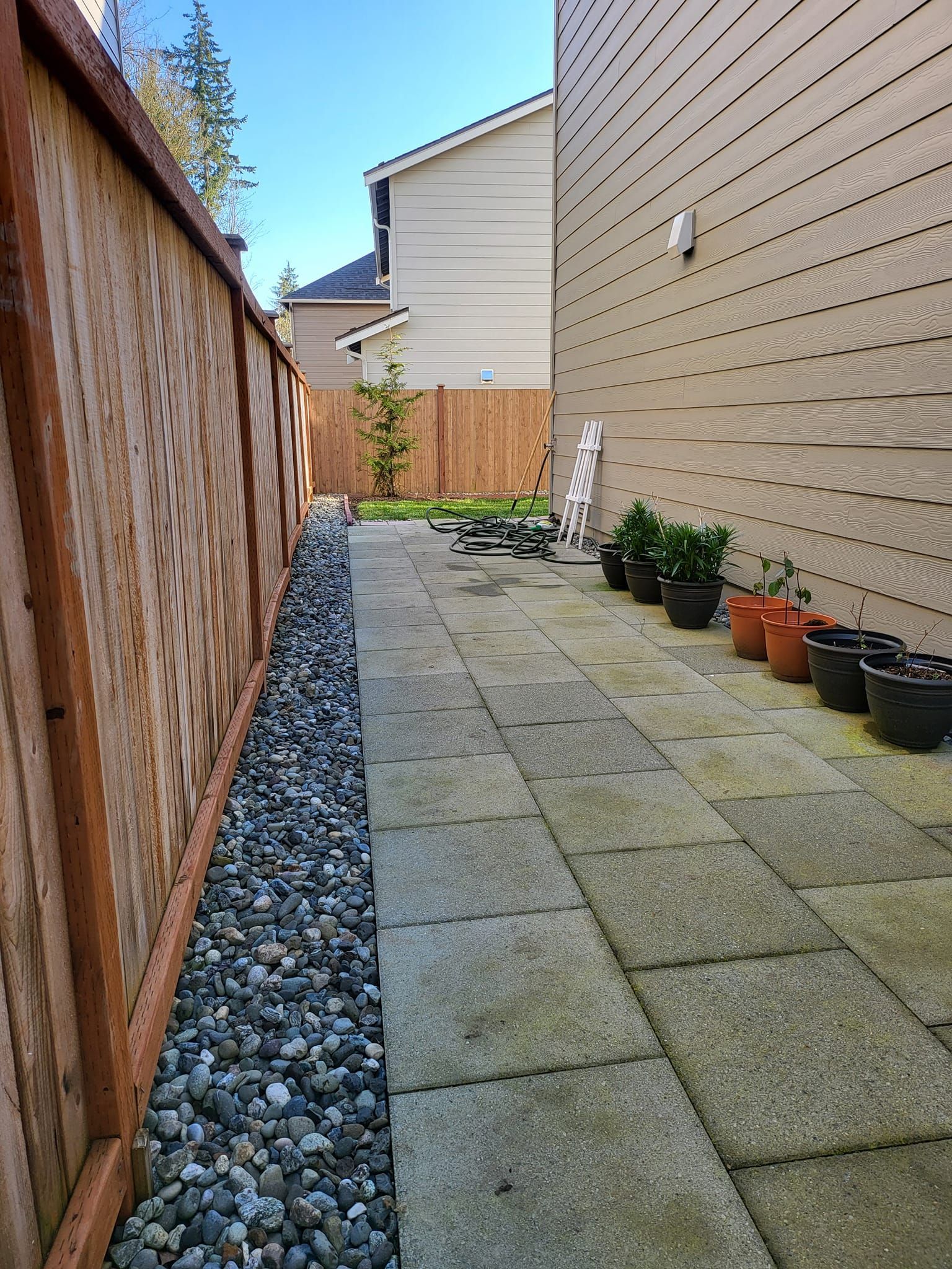 Narrow backyard walkway with stone pavers, gravel bed along a wooden fence, and potted plants.