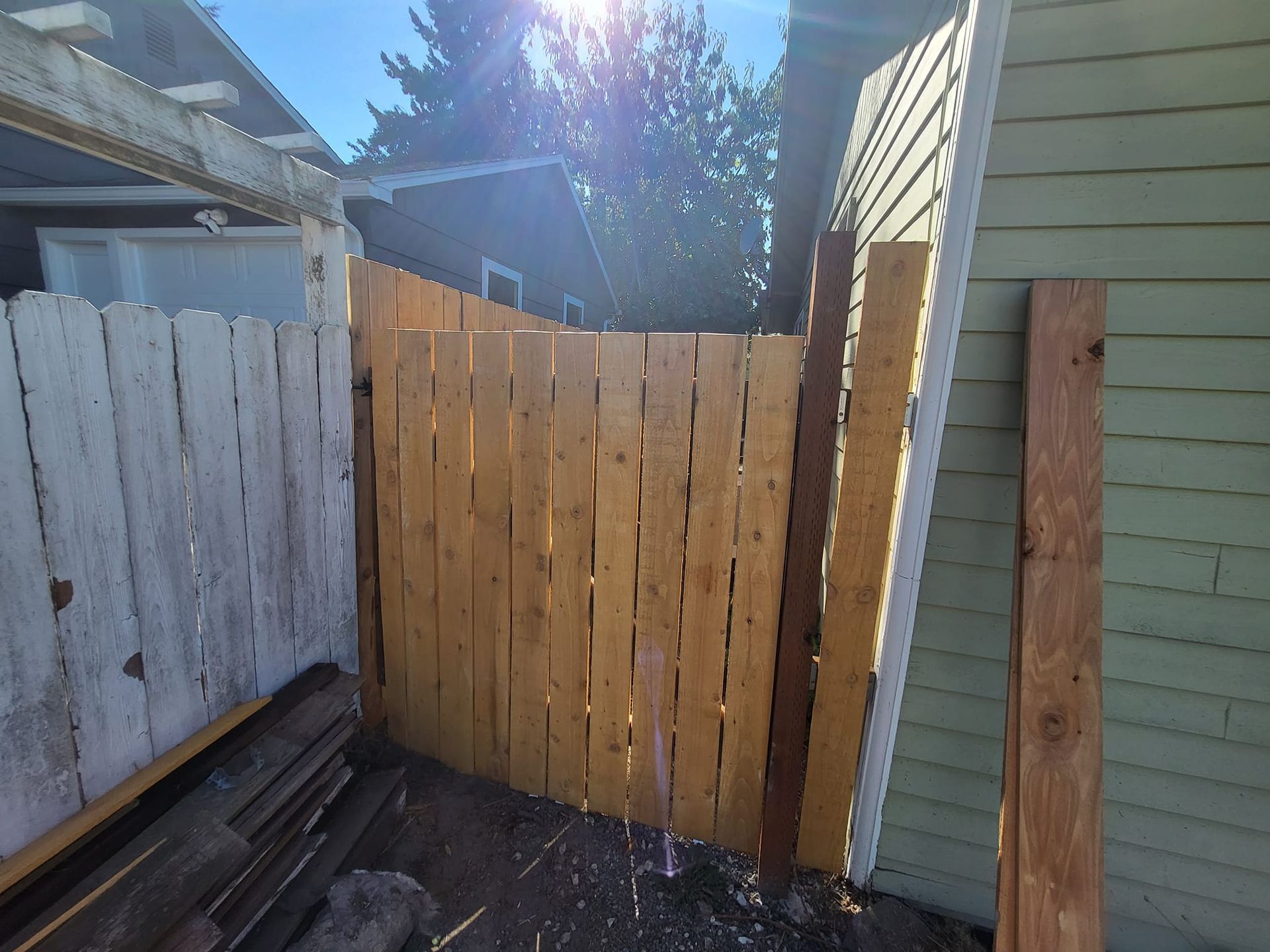 Wooden fence section between weathered white fence and green house siding; bright sun overhead.
