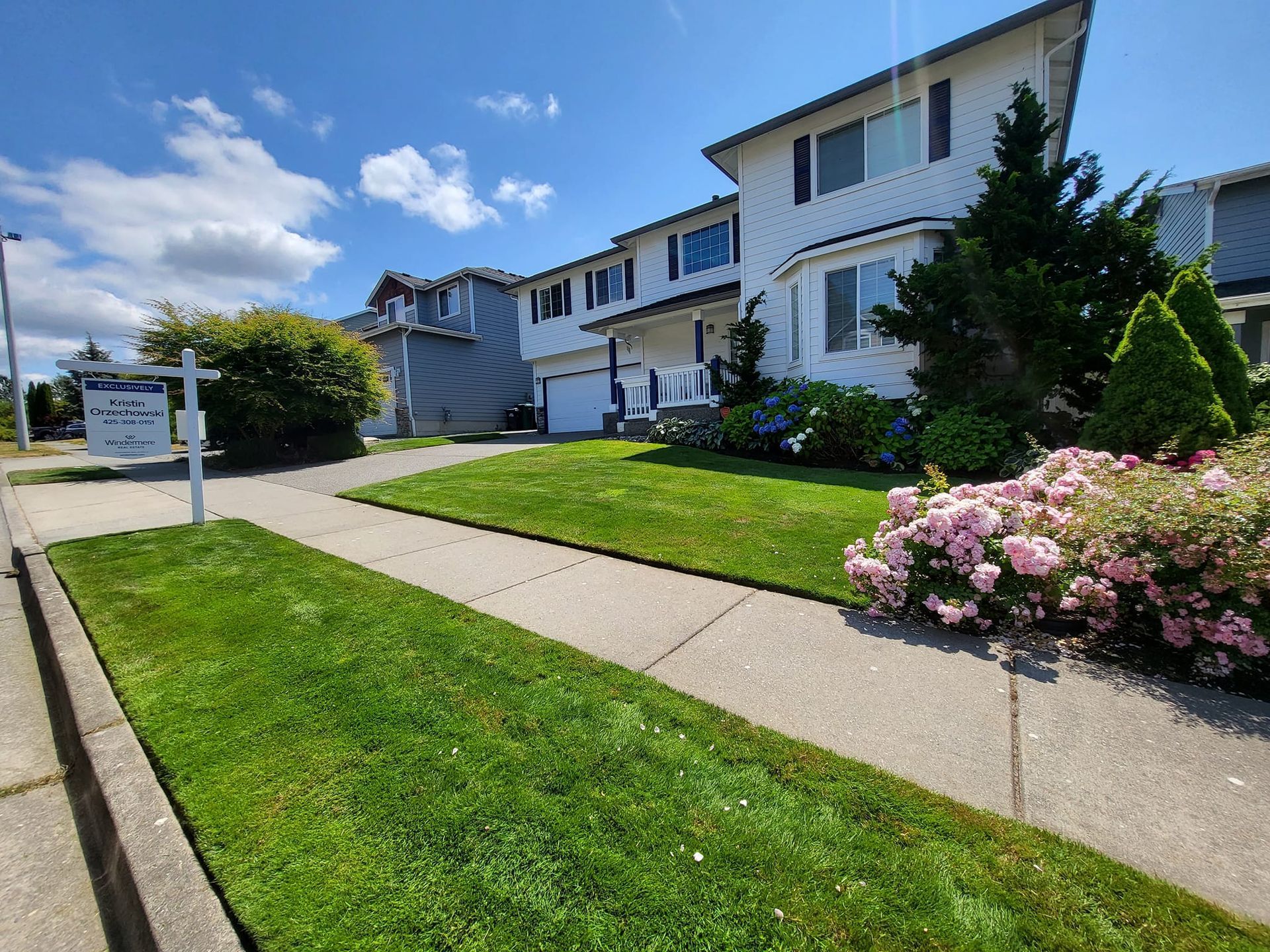 White house with green lawn and pink flowers, real estate sign on the sidewalk.