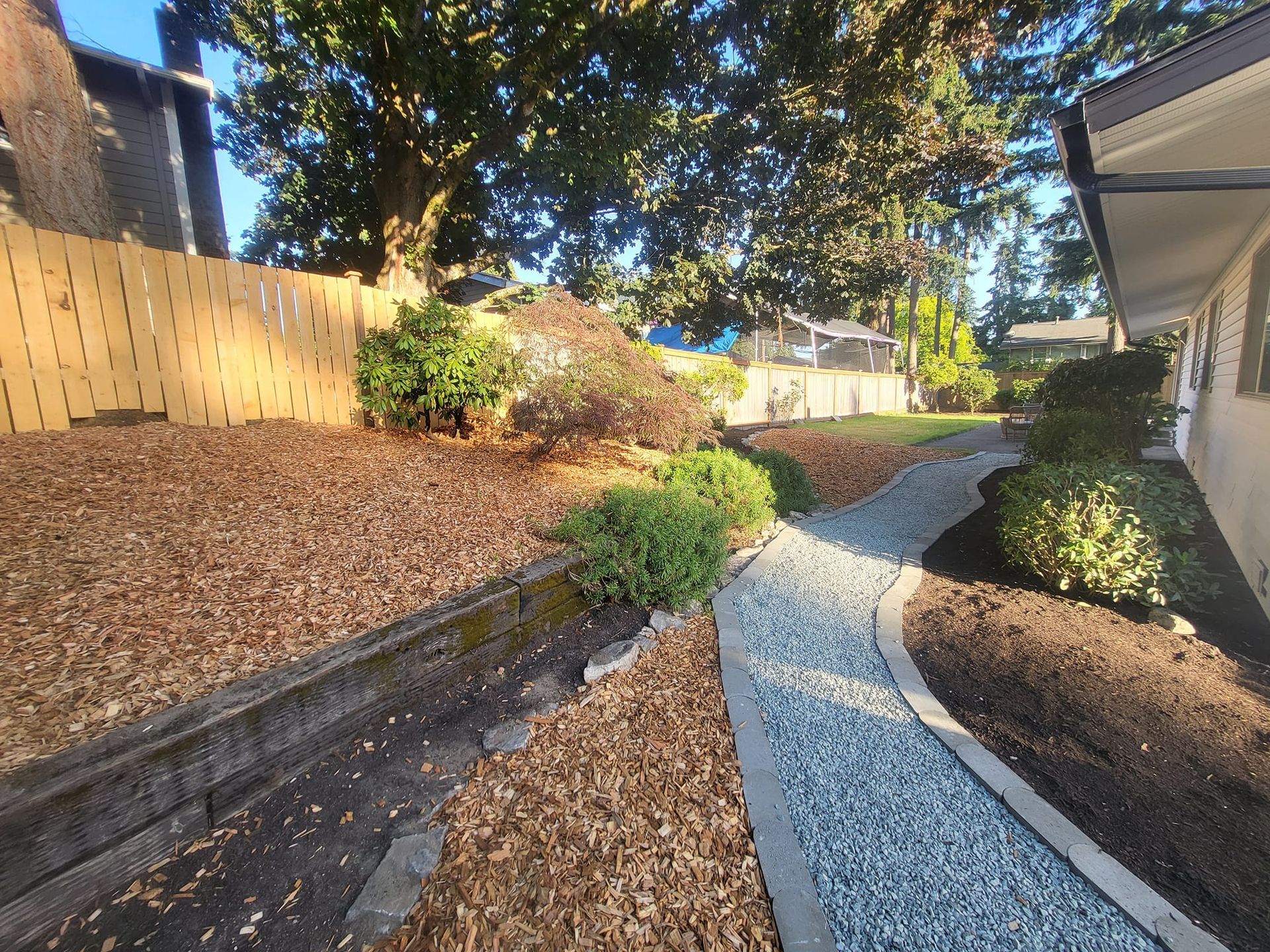 Landscaped yard with gravel pathway, mulch beds, and wooden fence. Sunny day.