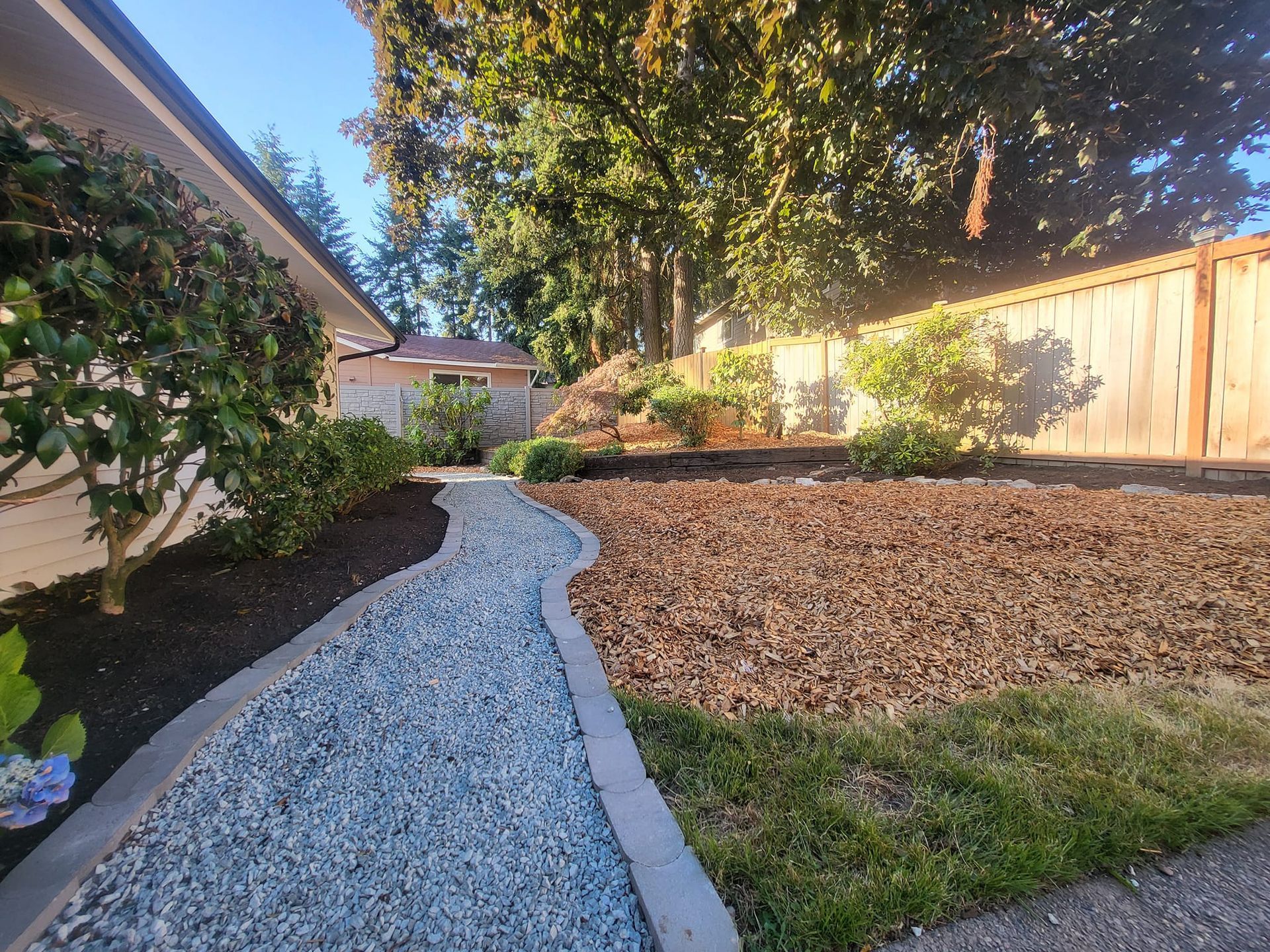 A gravel pathway leads from a home toward a landscaped yard with wood chip beds and a wooden fence.