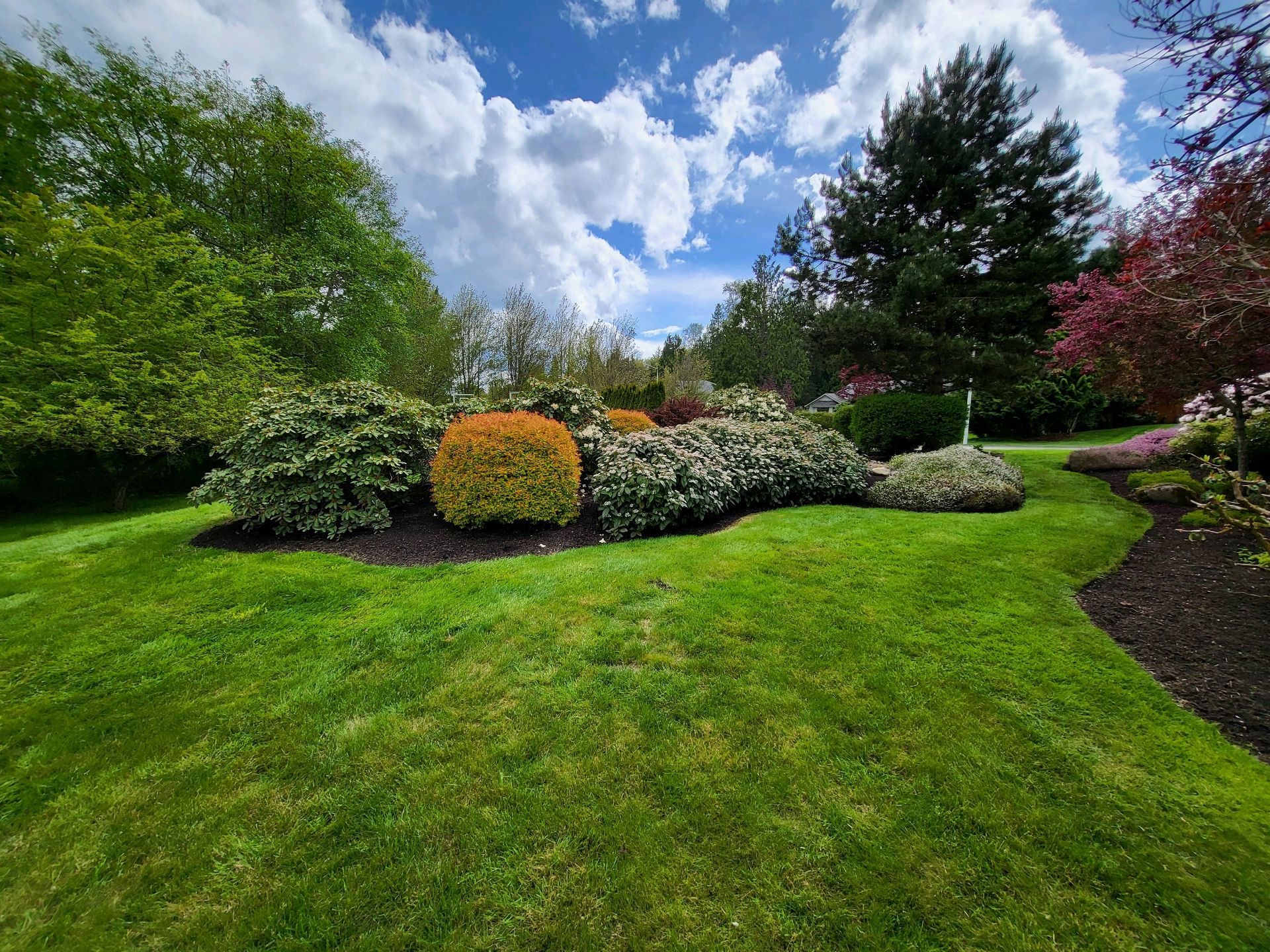 Lush green lawn with colorful flower beds, trees, and a partly cloudy blue sky.