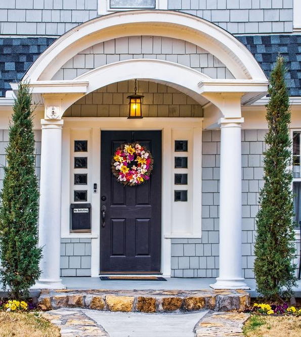 Front view of a house with a dark door, wreath, and arched portico. Two tall green trees flank the entrance.