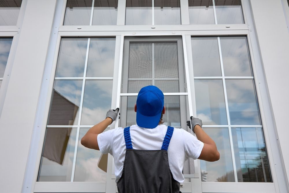 A worker in a white shirt and gray overalls installs a window screen into a large white-framed window.