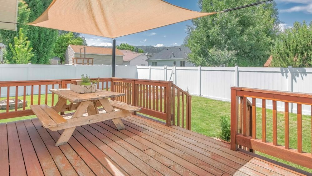 Wooden backyard deck with picnic table, beige umbrella, and white fence in sunny outdoor setting