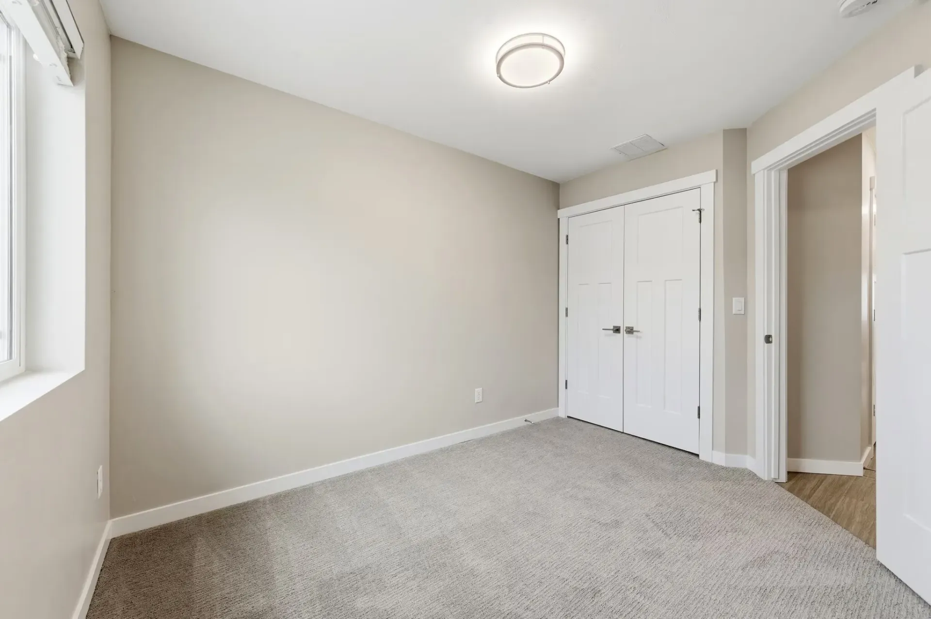 Interior bedroom with beige walls, light carpet, a window on the left, and double-door closet.