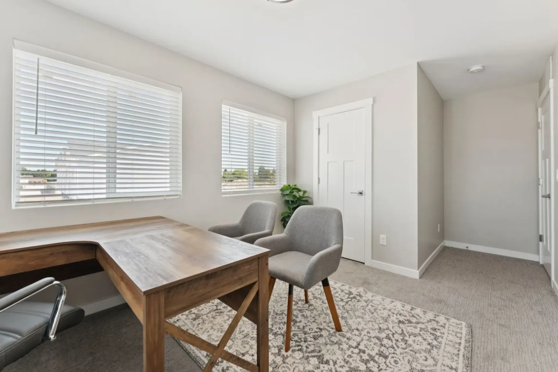 Bright home office in an apartment with a wooden L-shaped desk, two gray chairs, and large windows with blinds.