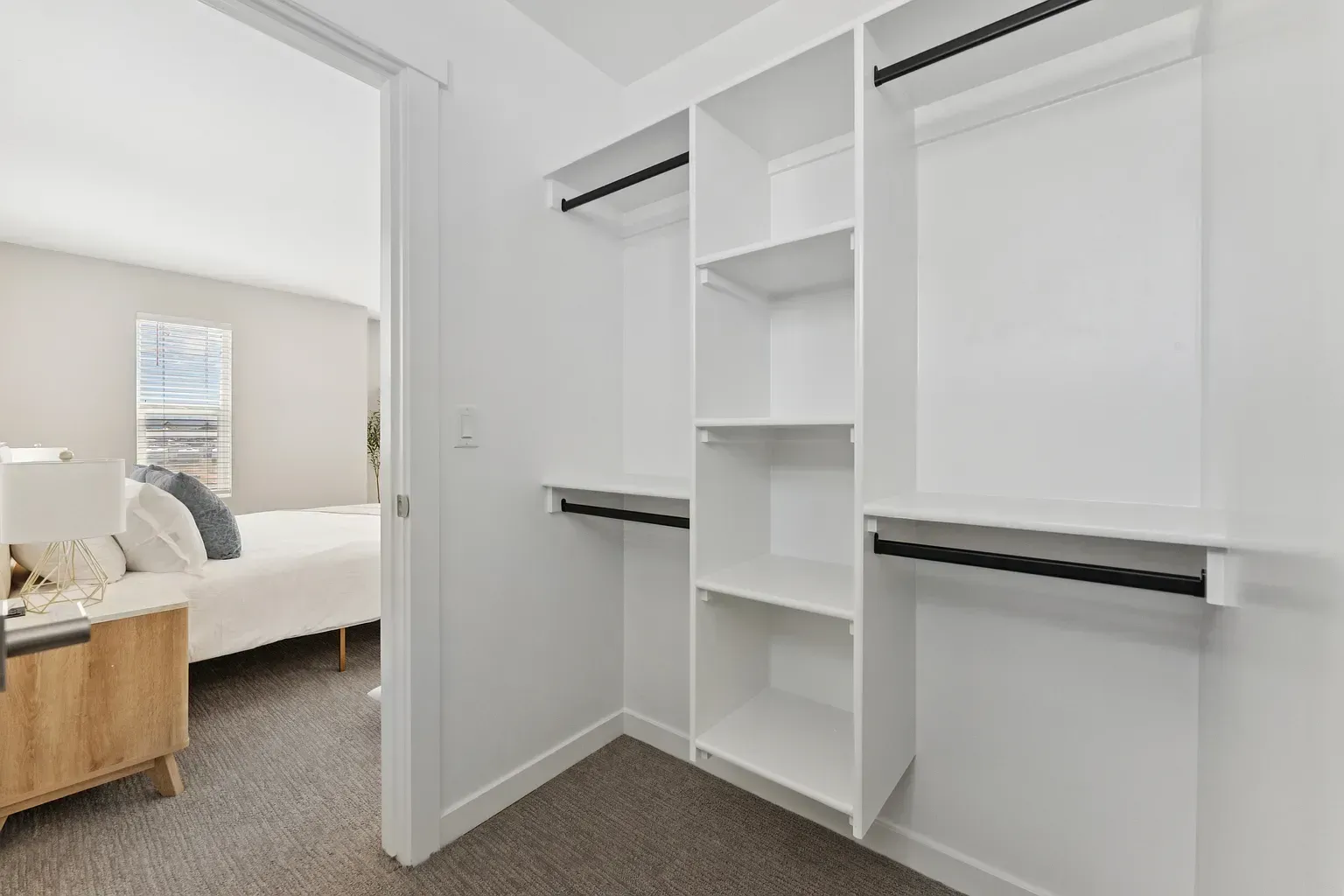Walk-in closet with white built-in shelves and hanging rods, adjacent to a bedroom.