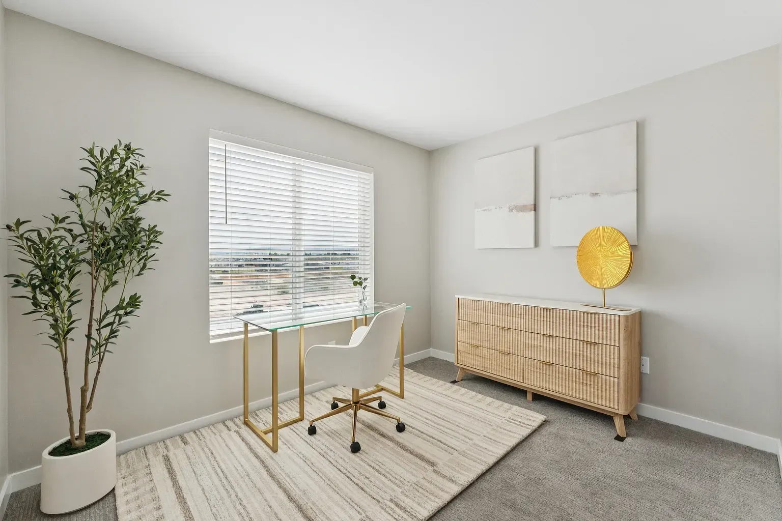 Bright apartment home office with glass desk, white chair, a tall plant, and a wooden dresser.