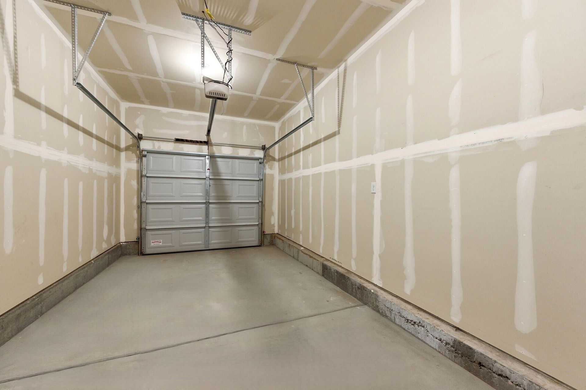 Empty residential garage with a metal roll-up door, exposed drywall seams, and a concrete floor.