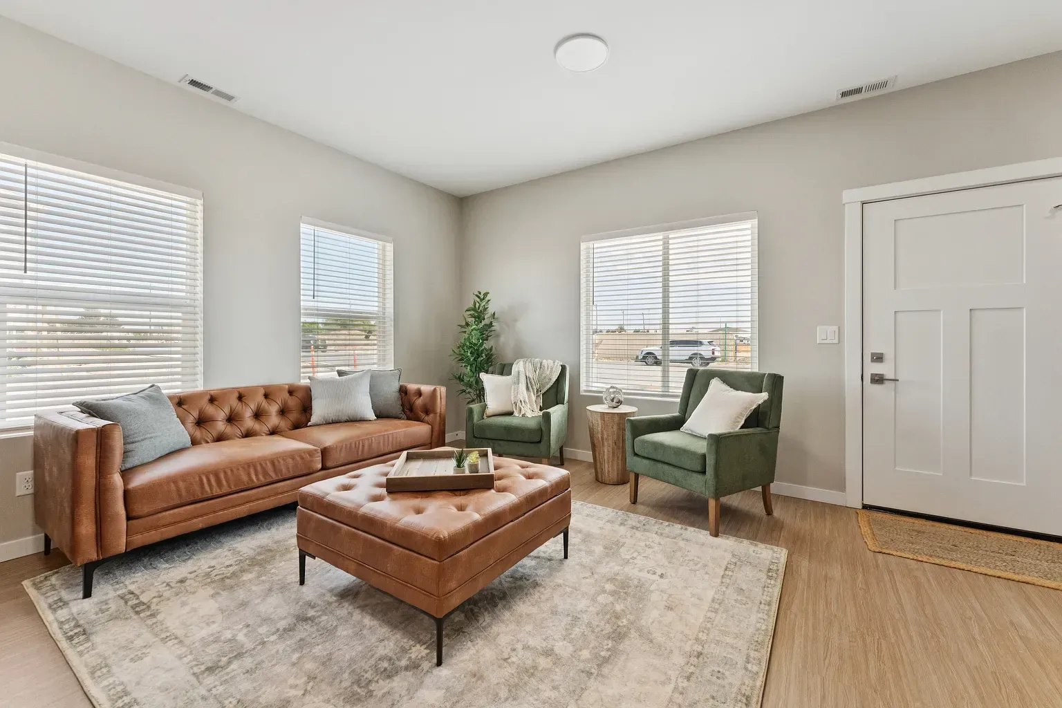 Sunlit living room with a tan tufted sofa, two green accent chairs, and a coffee ottoman.