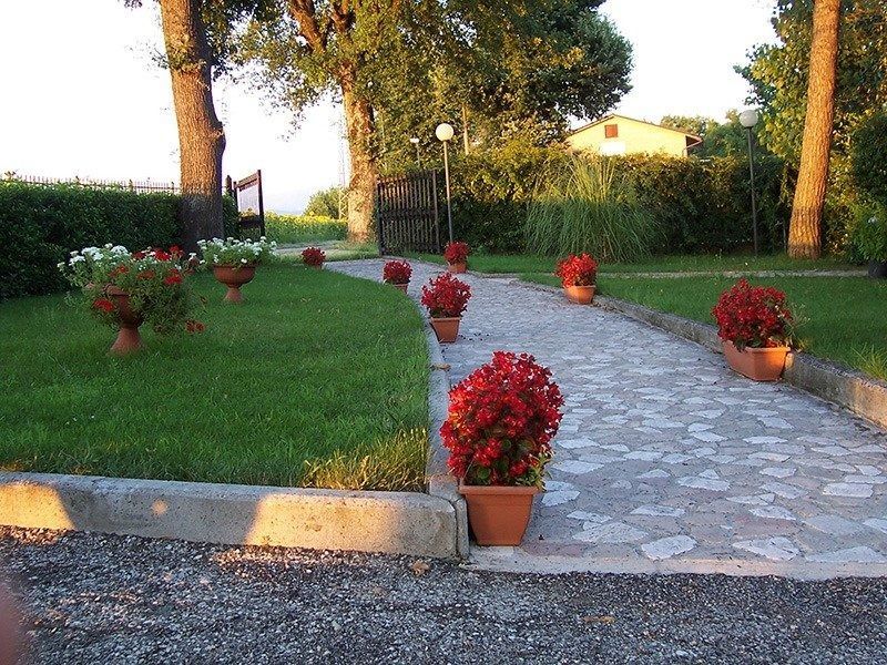 Sentiero del giardino fiancheggiato da vasi di fiori rossi, accanto a un prato verde e ad alberi al tramonto.