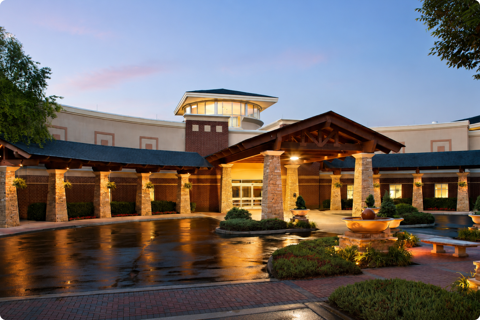 Illuminated lodge entrance with stone columns, wet courtyard, and landscaped gardens at dusk