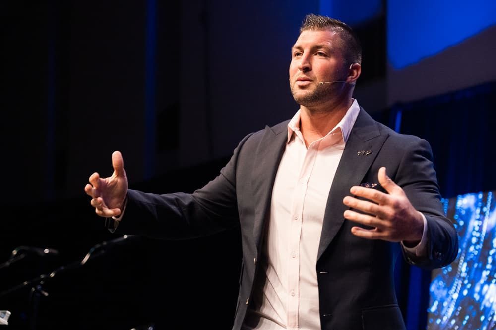 Man speaking on stage in a dark blazer and white shirt, gesturing with both hands under blue lighting