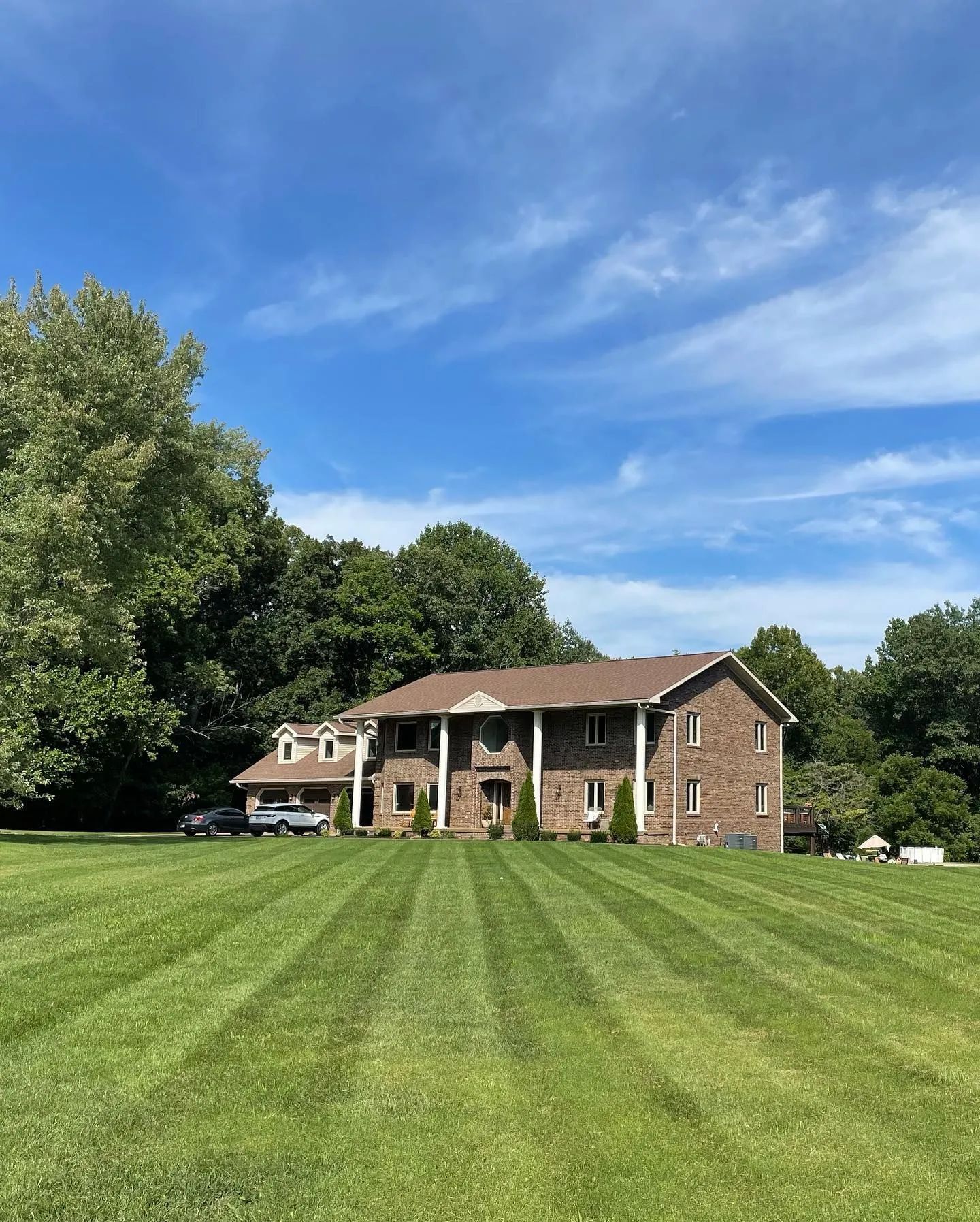 Large brick house with a wide striped lawn under a bright blue sky, framed by trees.