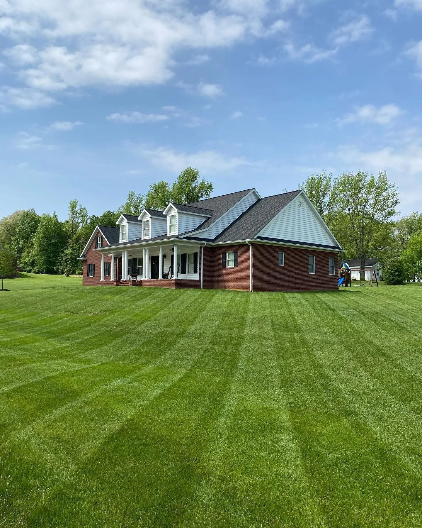 Red brick house with white porch and gray roof on a large striped green lawn under a blue sky