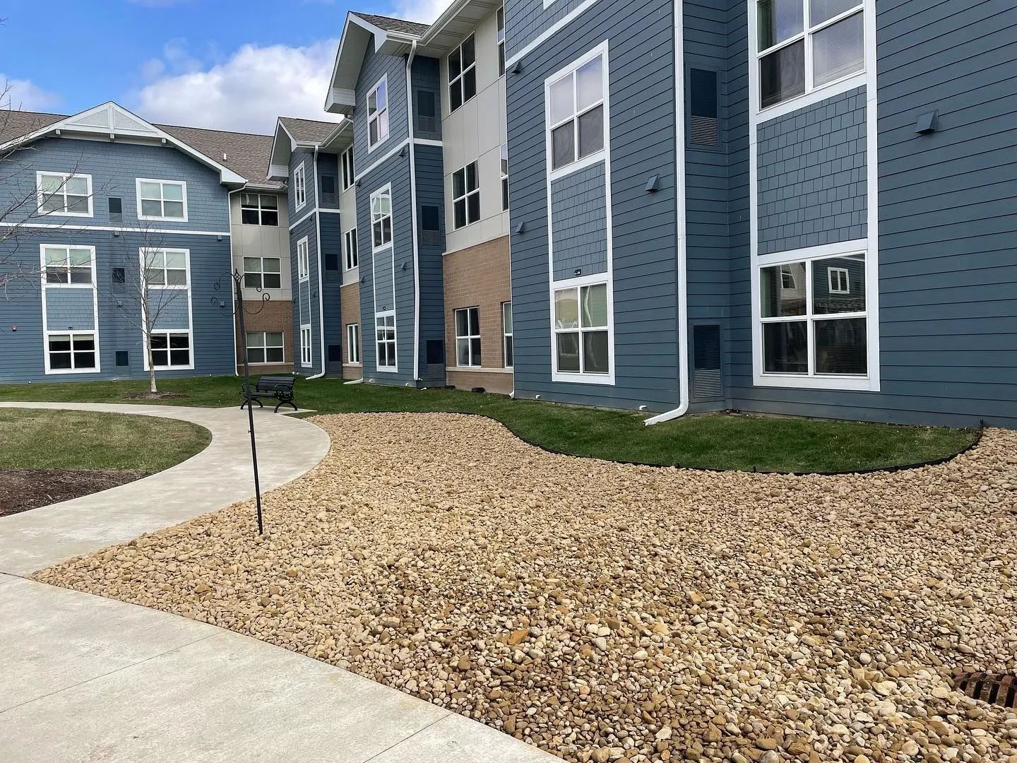 Blue apartment buildings beside a curving sidewalk and gravel landscape under a partly cloudy sky